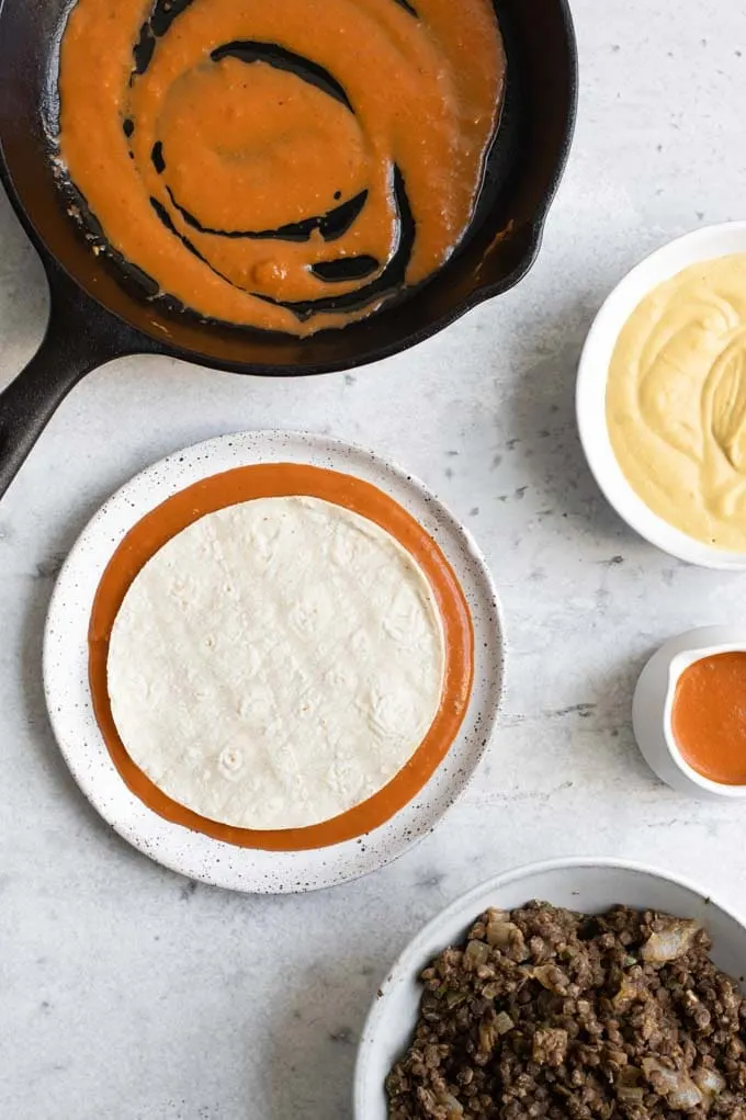 preparing the enchiladas, with everything laid out and a corn tortilla dipped in a plate of enchilada sauce