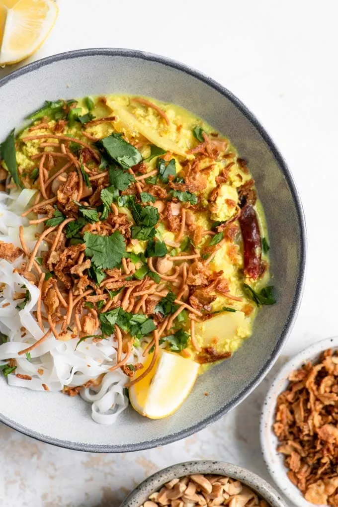close up of the kaukswe topped with crispy noodles, crispy onion, chopped roasted peanuts, minced chili, cilantro, and a wedge of lemon