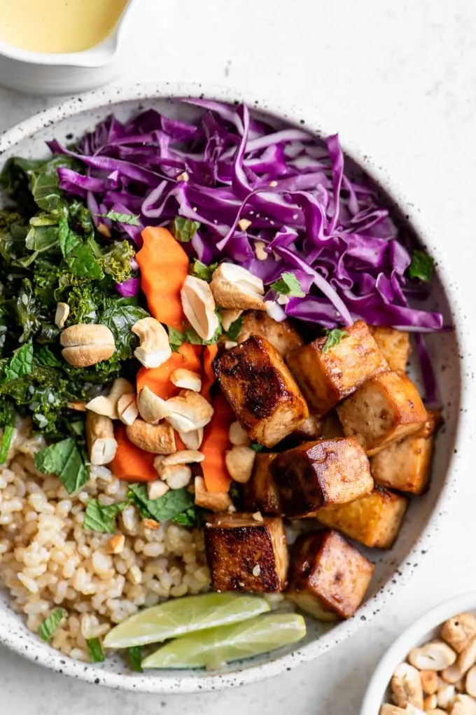 close up of the tofu buddha bowl with sesame kale, red cabbage, carrots, brown rice, toasted cashews and lime wedges
