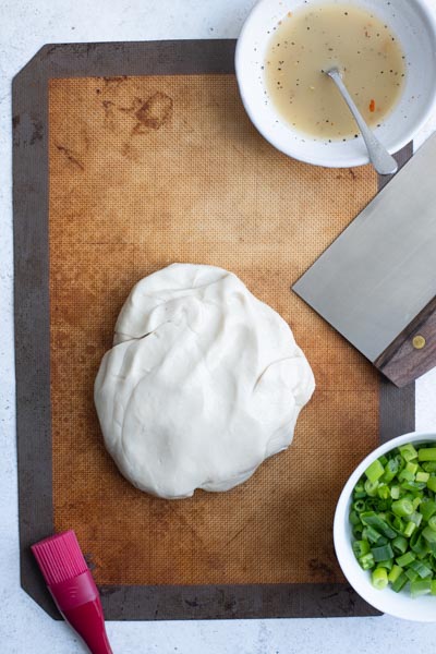 the scallion pancake dough on a silicon mat and a bowl of the oil-flour mixture and scallions