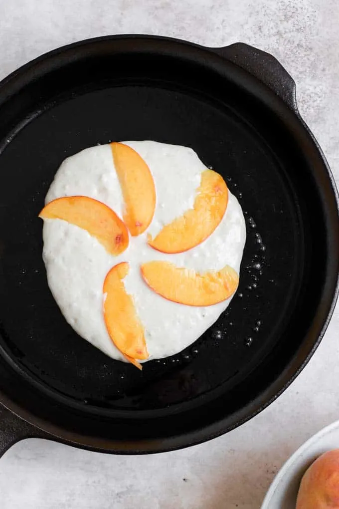 pancake on skillet with 5 slices of peach arranged on top in a fan pattern
