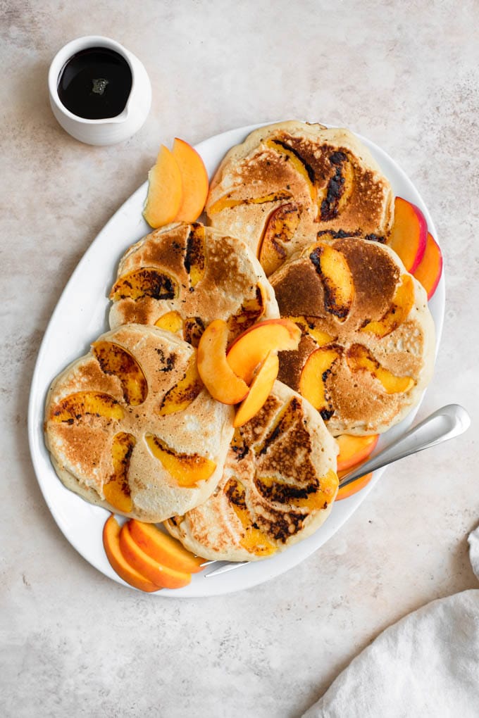 vegan peach pancakes on an oval platter with a small pourer of maple syrup on the side