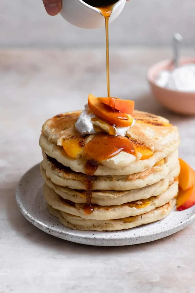 pouring maple syrup over a stack of peach pancakes