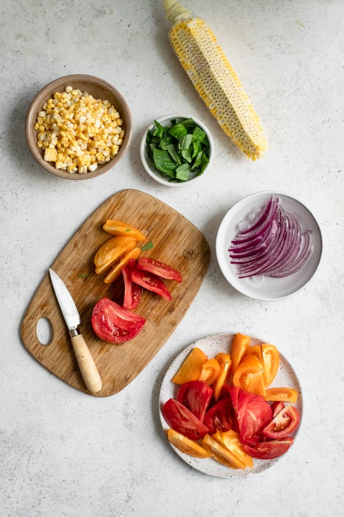 ingredients cut and prepared for salad, red onion in ice water, tomato being cut on cutting board, corn on the cob with kernels cut off, basil chopped