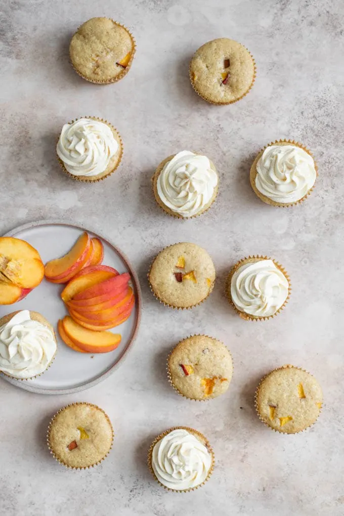 overhead photo of peach cupcakes, half of them frosted, with a plate of sliced peaches