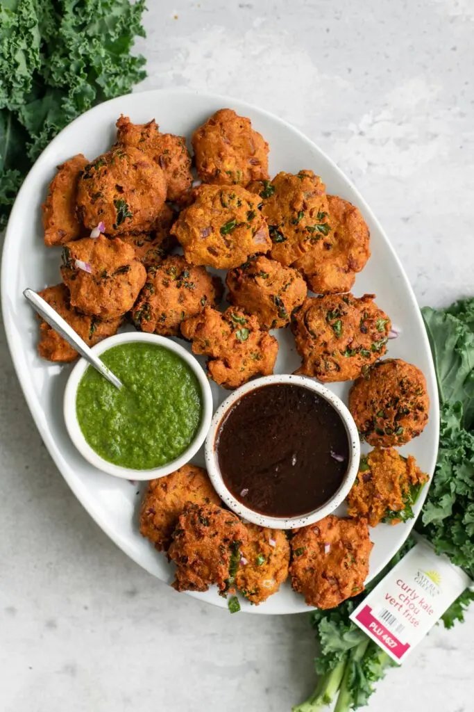 kale pakoras served with tamarind chutney and cilantro chutney with a bunch of nature's greens kale next to the plate