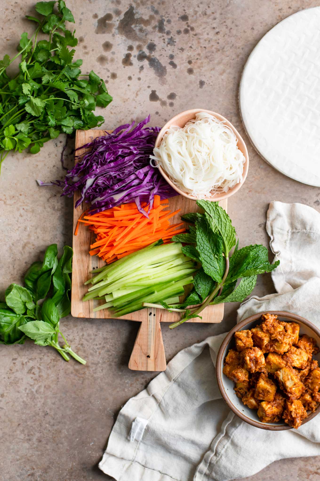 prepared ingredients for spring rolls set out: julienned red cabbage, carrots, cucumber, fresh basil, cilantro, mint, rice vermicelli, peanut tofu, and rice paper wraps