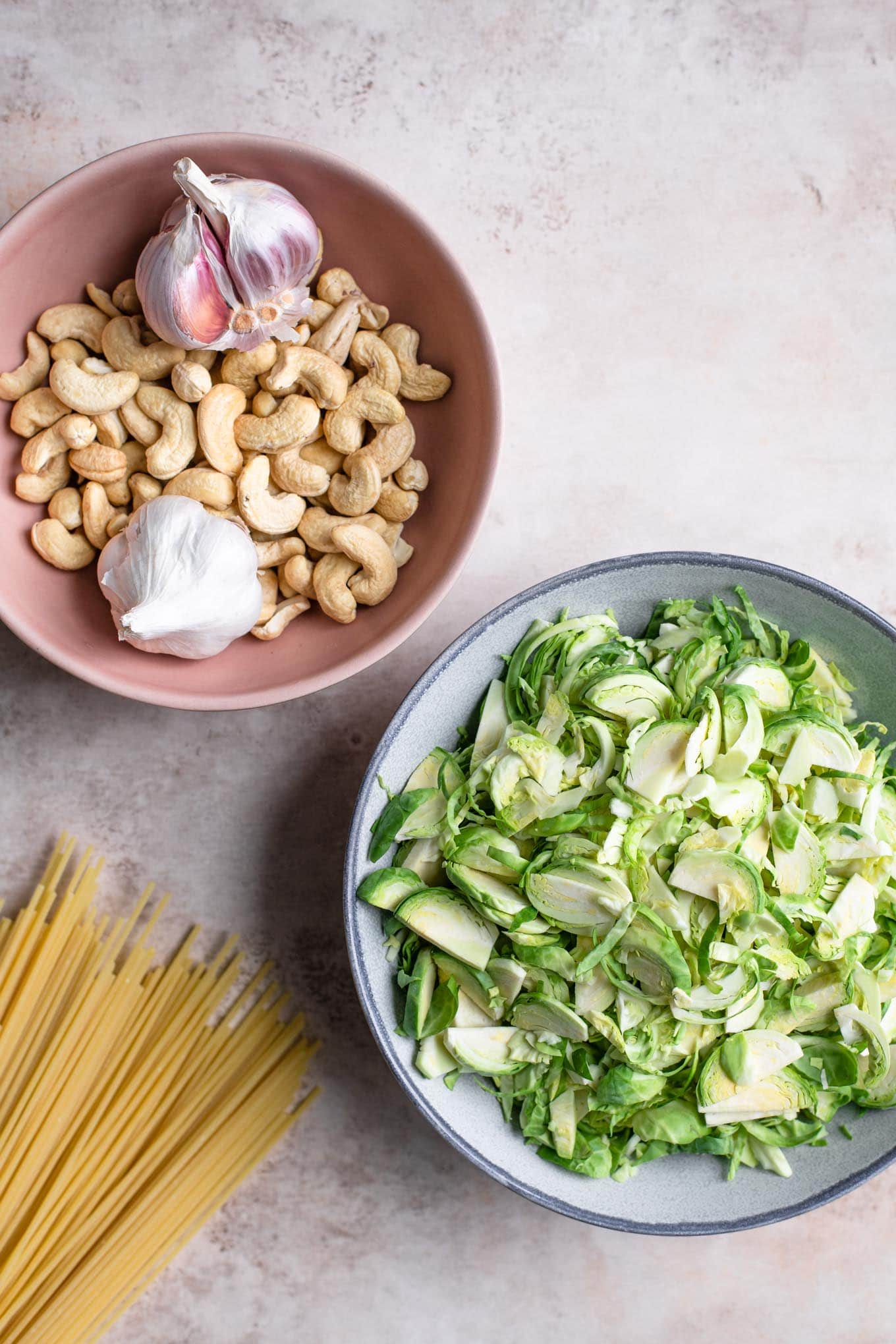 shaved brussels sprouts in a bowl and cashews and garlic in a second bowl with dry spaghetti spread out on the counter