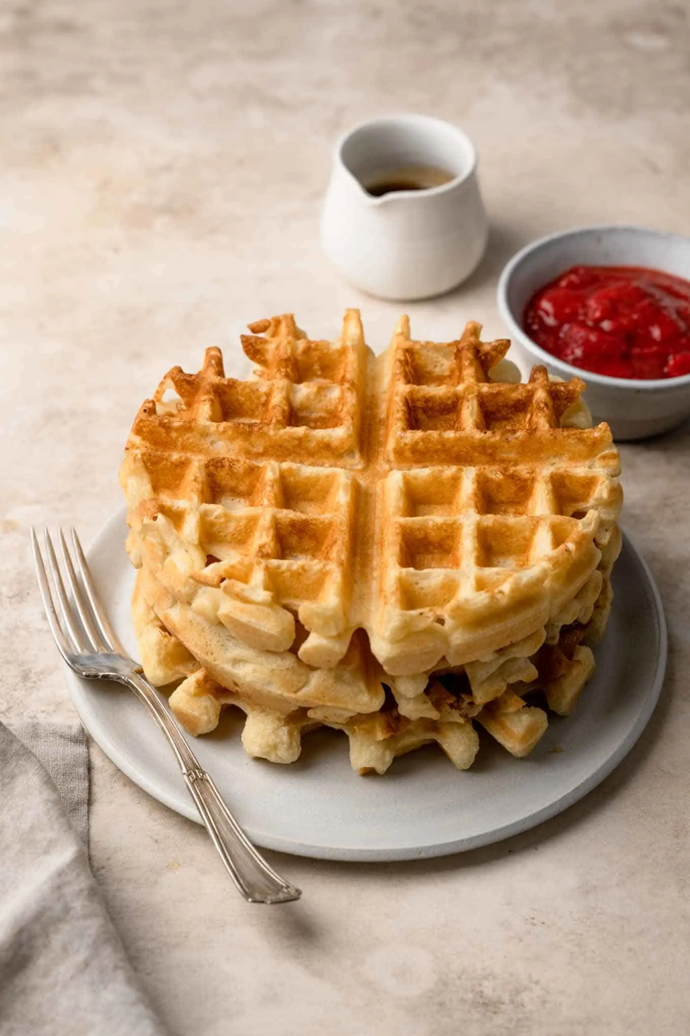 Three waffles stacked on a plate with a jug of maple syrup and fruit compote to serve
